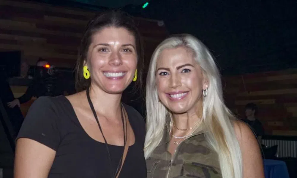 Two smiling women posing indoors, one with long blonde hair in camouflage top, the other with dark hair and yellow earrings.