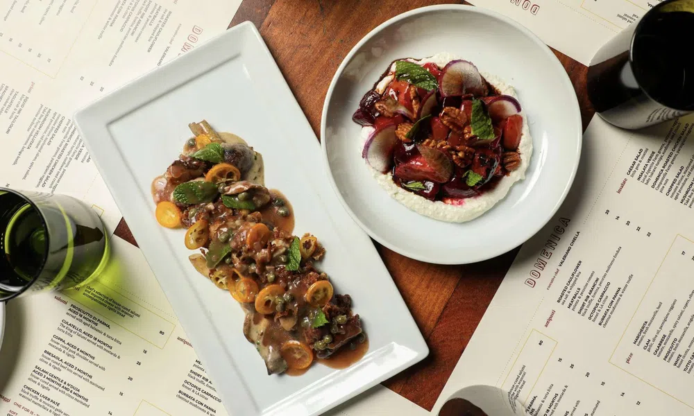 Gourmet dishes with sauces and herbs served on white plates surrounded by menus and wine glasses on wooden table.