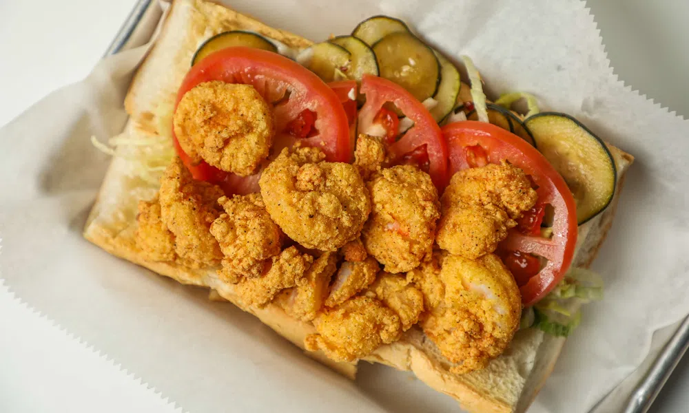 Fried shrimp po'boy sandwich with sliced tomatoes, pickles, and lettuce on a sub roll.