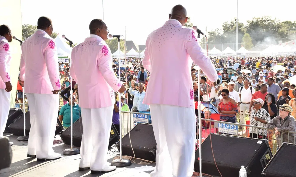 Five male singers in pink jackets performing on outdoor stage before large audience at daytime event.