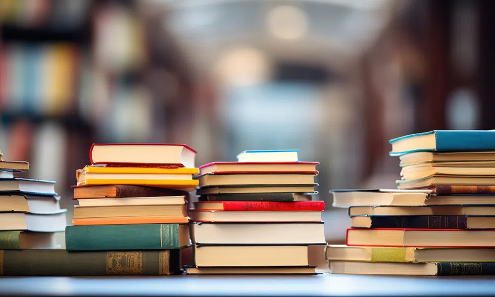 Stacks of colorful hardcover books arranged on a table in a blurred library setting with warm lighting.
