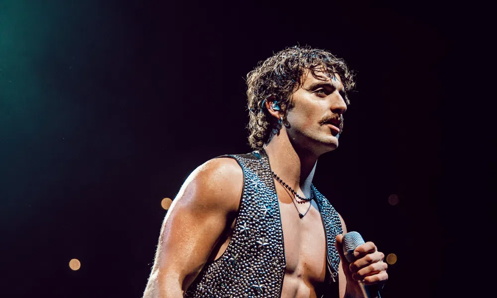 Muscular male singer with curly hair and sleeveless studded vest performing on stage with microphone