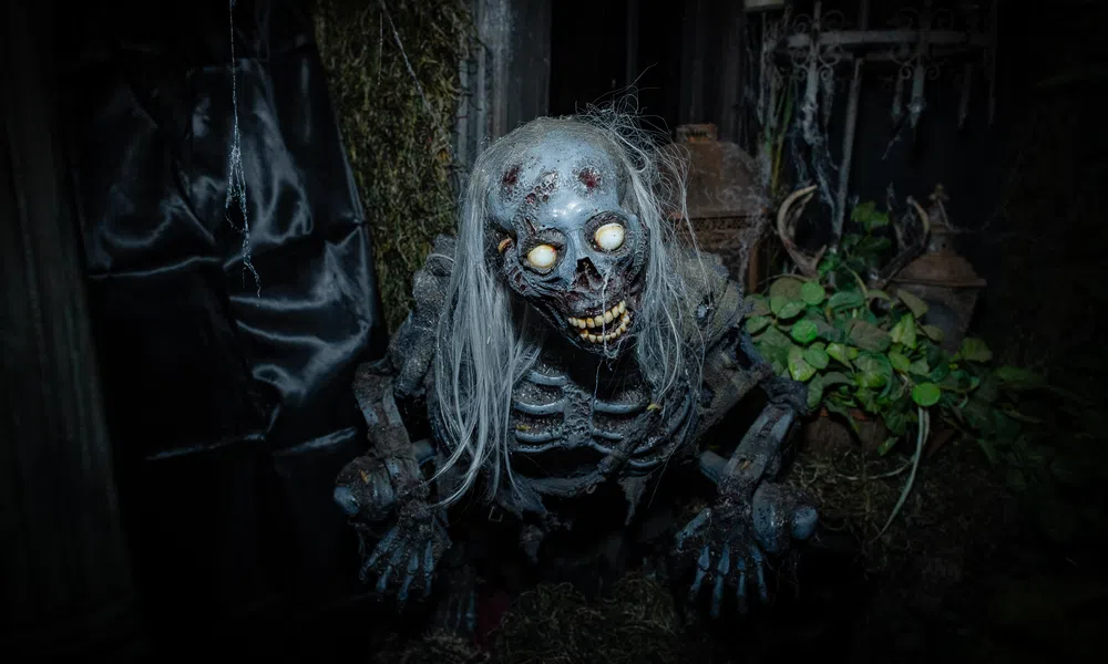 Creepy skeleton figure with glowing eyes and long white hair in a dark, eerie setting with plants and moss.