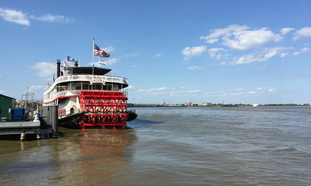Red and white paddlewheel riverboat docked on a wide river under a blue sky with scattered clouds.