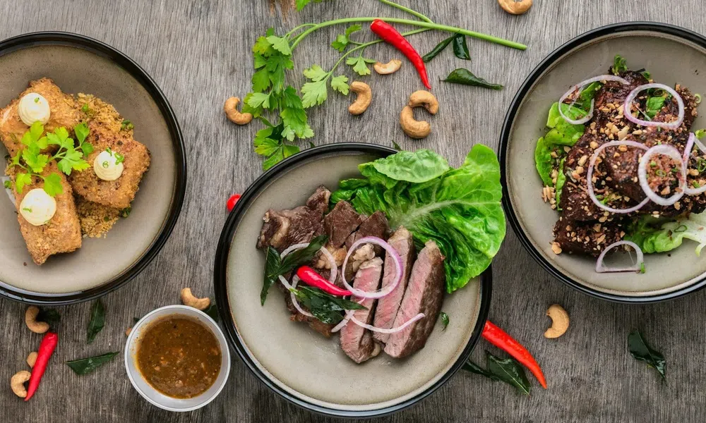 Three plates of gourmet dishes with grilled meat, fried appetizers, fresh herbs, lettuce, red chili, and cashew nuts on wooden table.