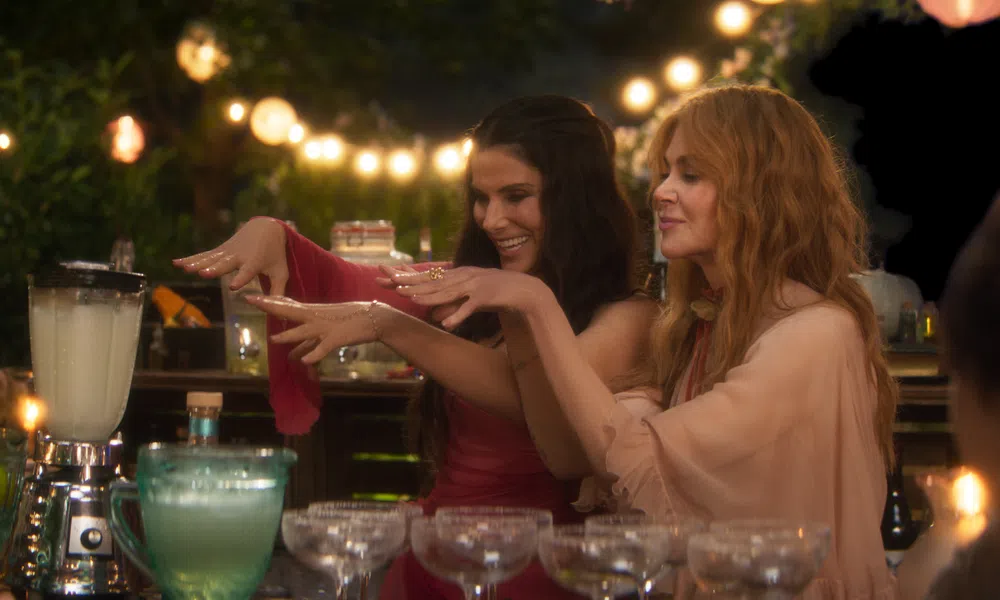 Two women at a festive outdoor bar preparing drinks with string lights and glassware in the background.