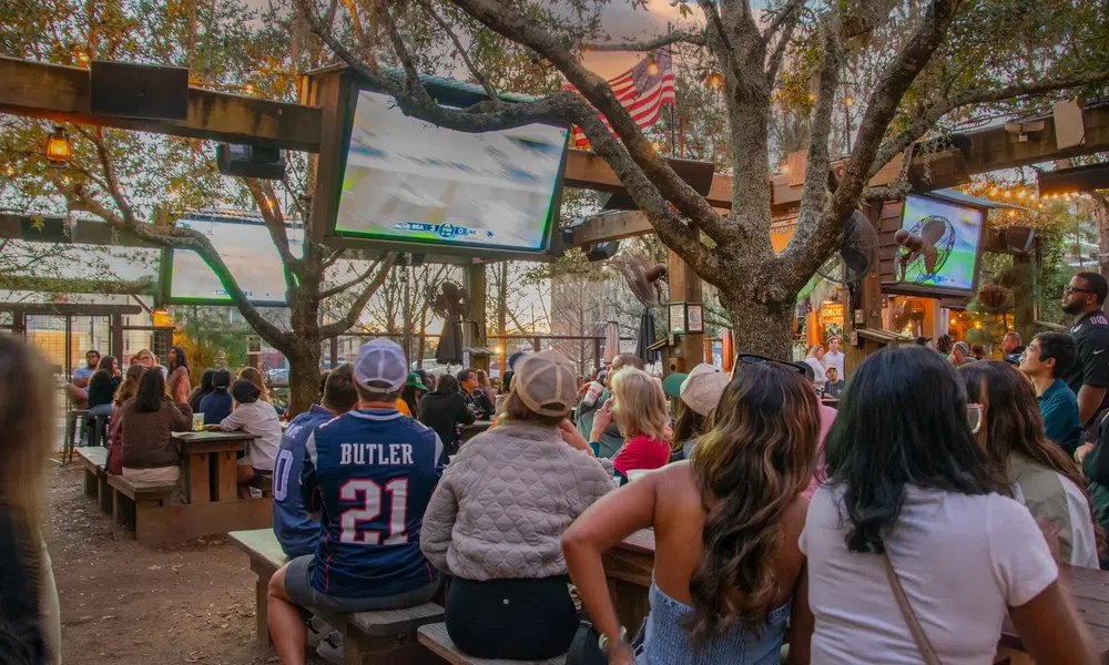 Outdoor sports bar with crowd watching a football game on multiple large screens under trees and string lights.