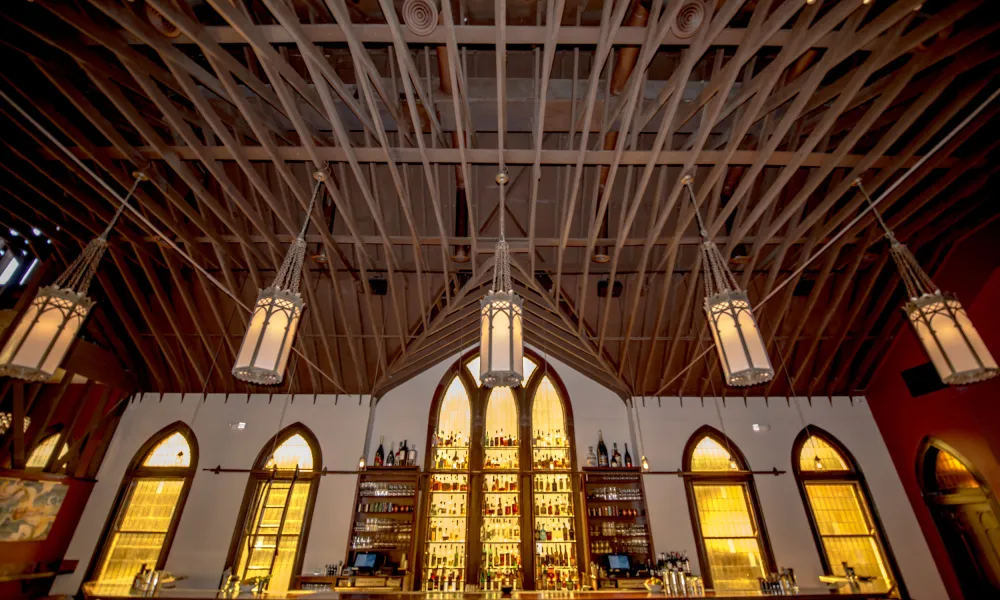 Upscale bar with gothic arched windows, illuminated shelves of liquor, and wooden ceiling beams with hanging lanterns.