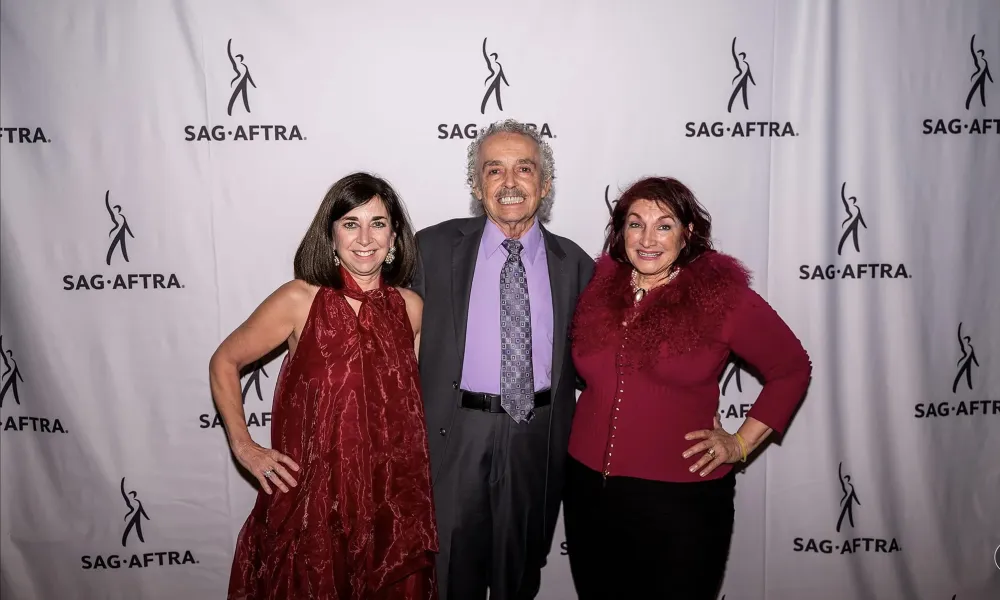 Three adults posing at SAG-AFTRA event with branded backdrop, two women in red outfits and one man in suit.