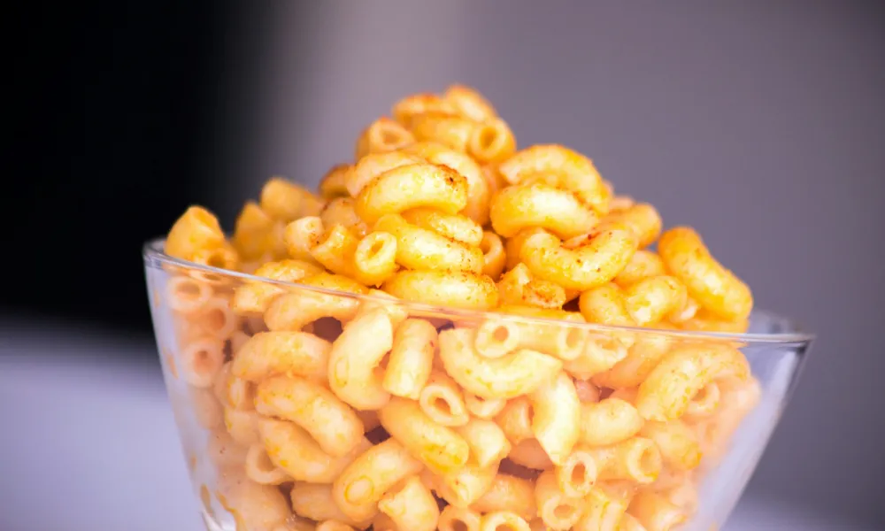Clear glass bowl filled with curly, orange seasoned cheese snacks on a white surface with blurred background