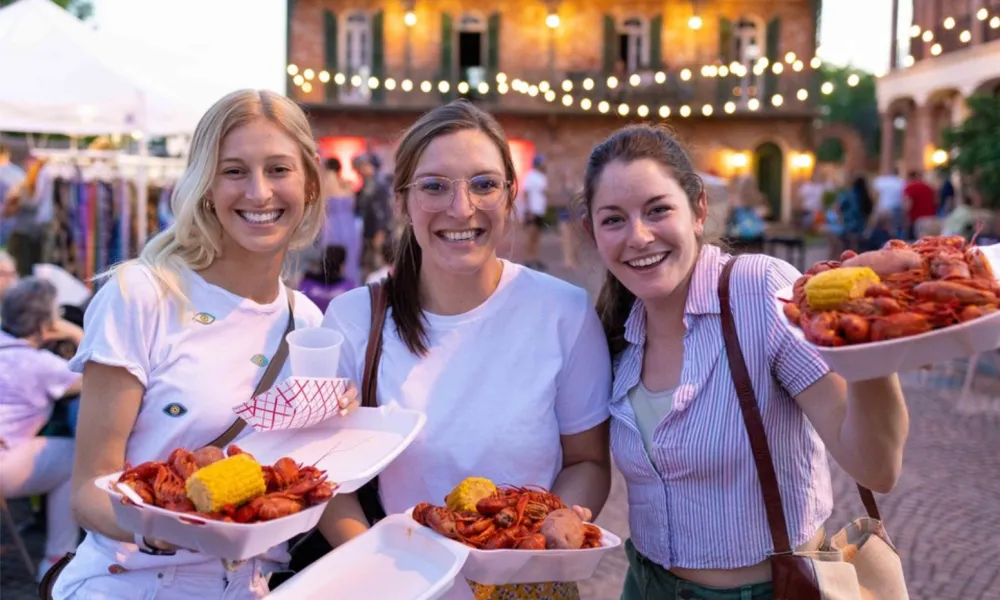 Three women holding trays of crawfish at an outdoor festival, smiling and enjoying the food.