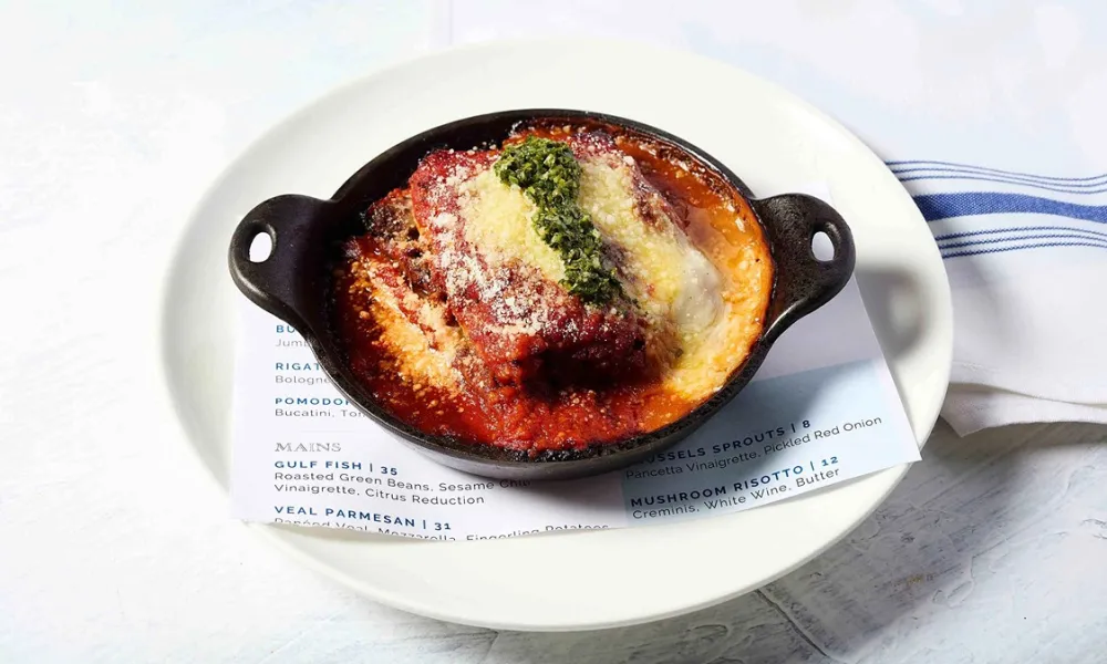 Baked veal parmesan with tomato sauce, cheese, and pesto served in a black dish on a white plate