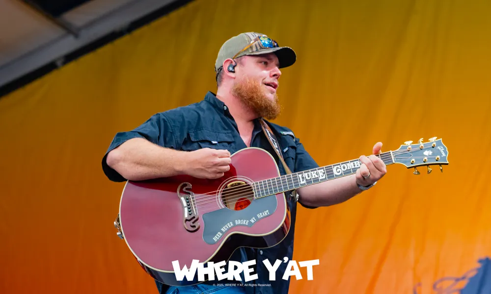 Male country musician playing guitar on stage with a yellow backdrop and wearing a cap and sunglasses
