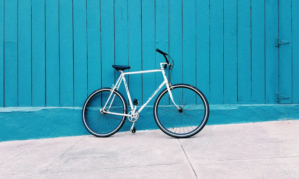 White bicycle parked against a vibrant blue wooden wall on a concrete pavement under sunlight.