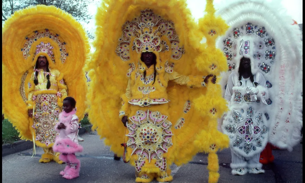 Three people in elaborate yellow and white beaded Mardi Gras costumes with large feathered headdresses stand outdoors