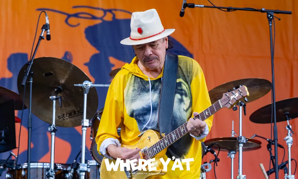 Guitarist wearing yellow hoodie and white hat playing electric guitar on stage with drums behind him