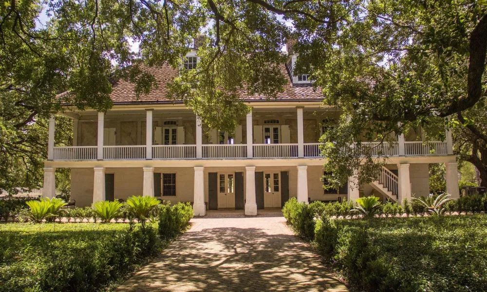 Historic two-story plantation house surrounded by lush greenery and shaded by large oak trees.