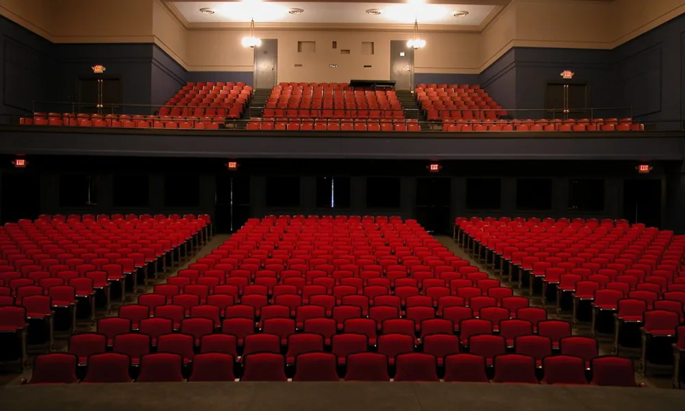 Empty theater with rows of red seats and a balcony, viewed from the stage, under warm lighting.