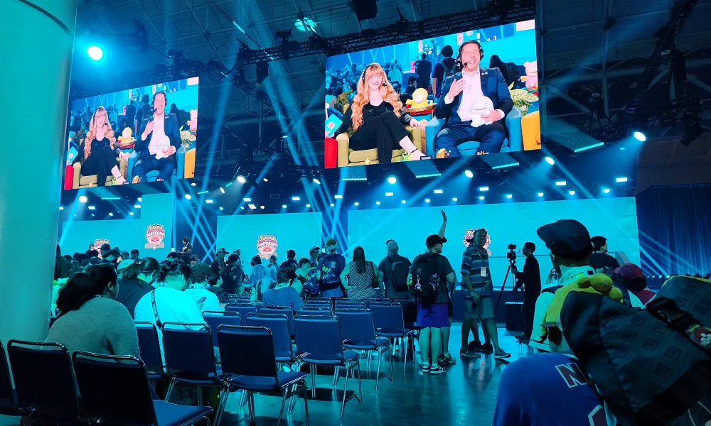 Crowded gaming event with audience watching live commentators on large screens under blue stage lighting.