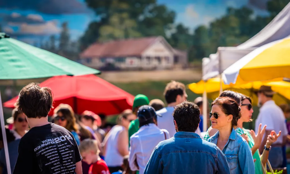 People socializing outdoors at a colorful market with red, green, and yellow umbrellas on a sunny day.