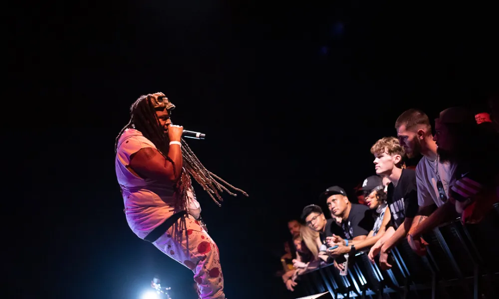Rapper with long dreadlocks performing on stage with microphone, engaging an attentive crowd in front row