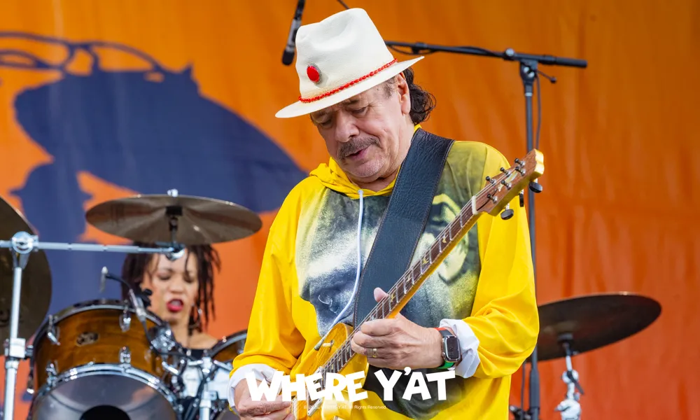 Guitarist in a white hat and yellow hoodie playing on stage with drummer in background against an orange backdrop.