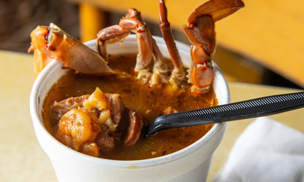 Close-up of a seafood stew with crab legs and shrimp served in a white foam cup with a black spoon.