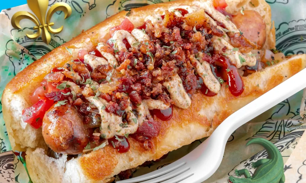 Loaded hot dog topped with crispy bacon bits, sauces, and diced tomatoes in a soft bun on patterned paper.