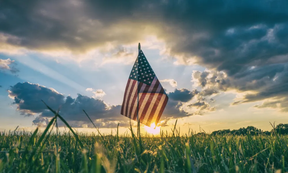 American flag standing in a grassy field with sun rays and dramatic cloudy sky at sunset