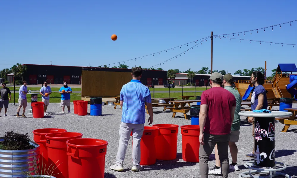 Group of men playing outdoor game tossing balls into large red bins on a sunny day with picnic tables and playground nearby