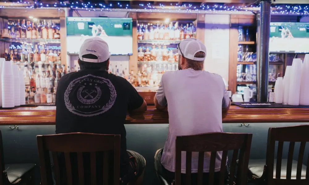 Two men wearing white caps watching a sports game at a bar with shelves of liquor and string lights.