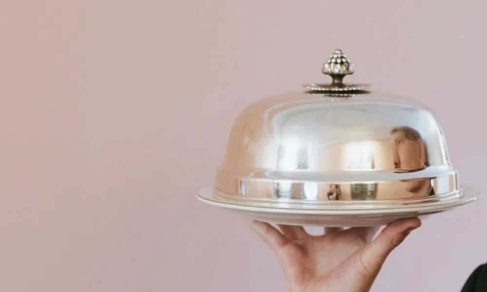 Person holding a silver cloche food cover on a tray against a soft pink background