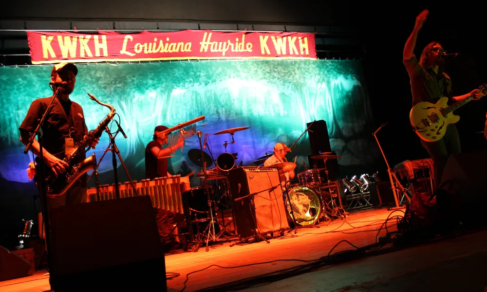 Live band performing on stage under KWKH Louisiana Hayride banner with saxophone, drums, and guitar.