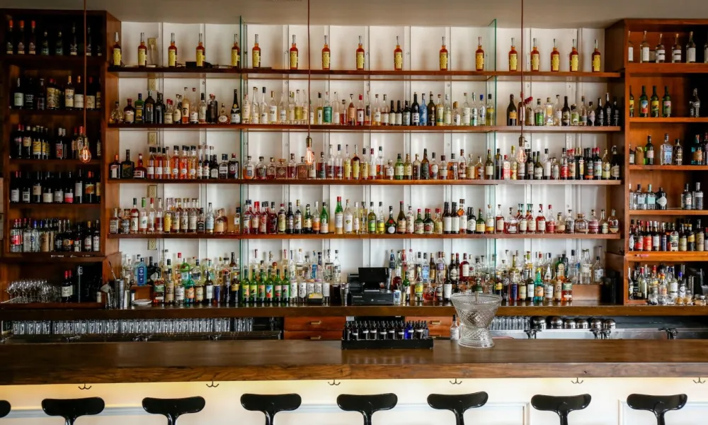 Wide bar with rows of liquor bottles on shelves and empty black bar stools lined up at wooden counter.