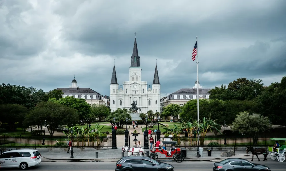 St. Louis Cathedral with horse-drawn carriages and cars on street under cloudy sky in New Orleans