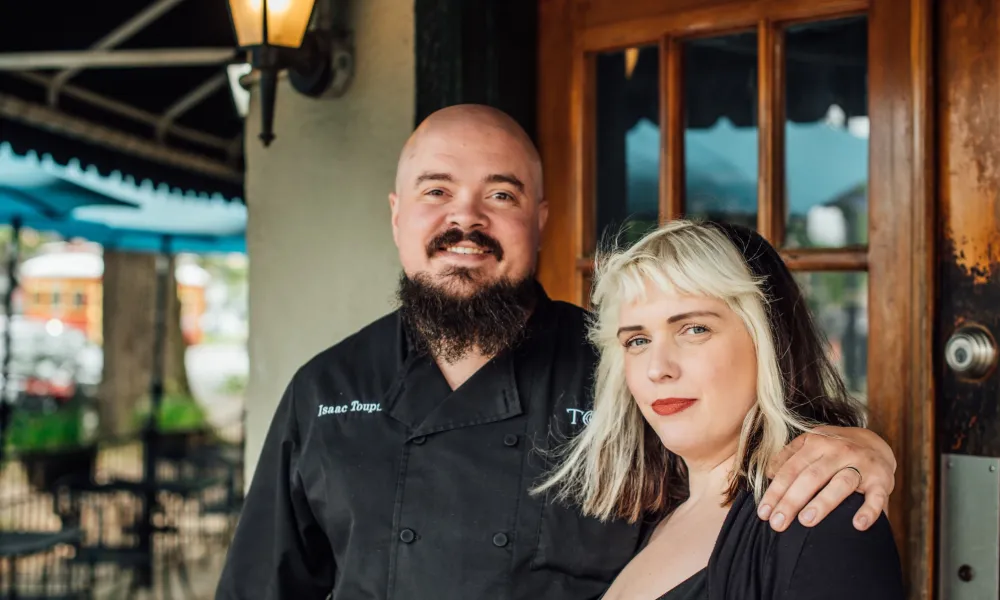 Smiling chef with beard in black uniform embraces woman with blonde hair outside restaurant entrance.