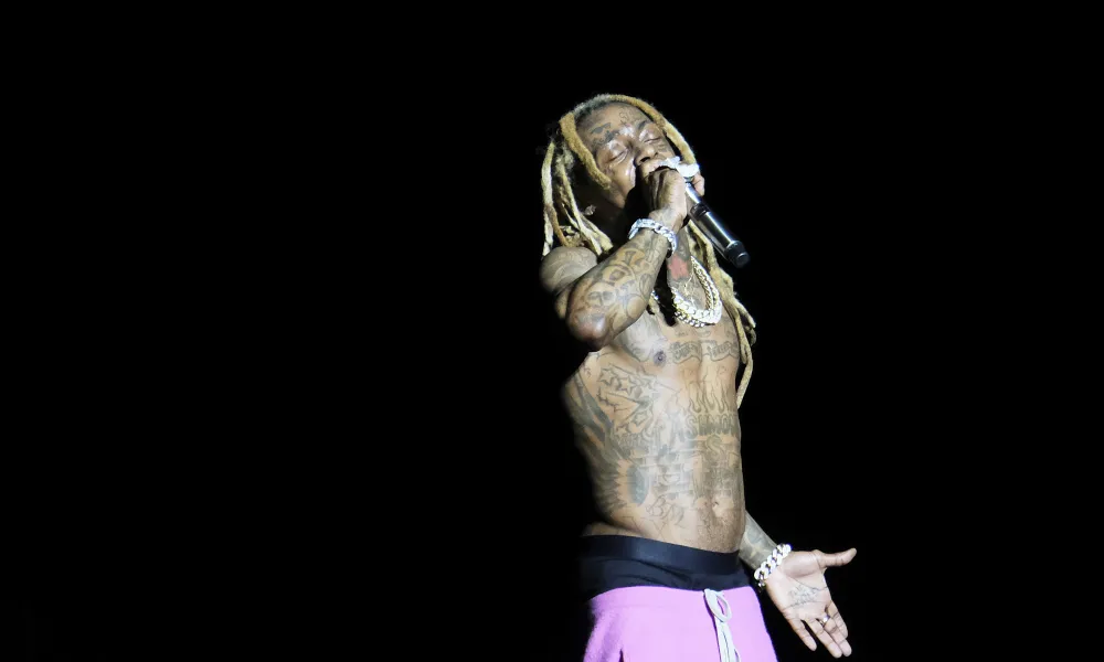 A tattooed rapper performs passionately on stage with a microphone, spotlighted against a dark background.