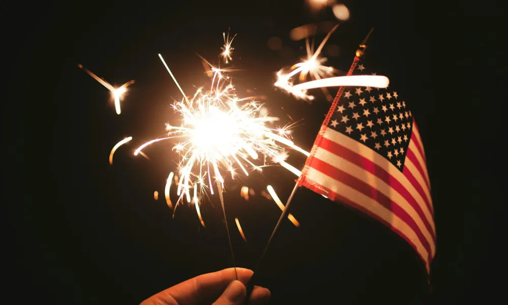 Hand holding a lit sparkler and a small American flag against a dark background