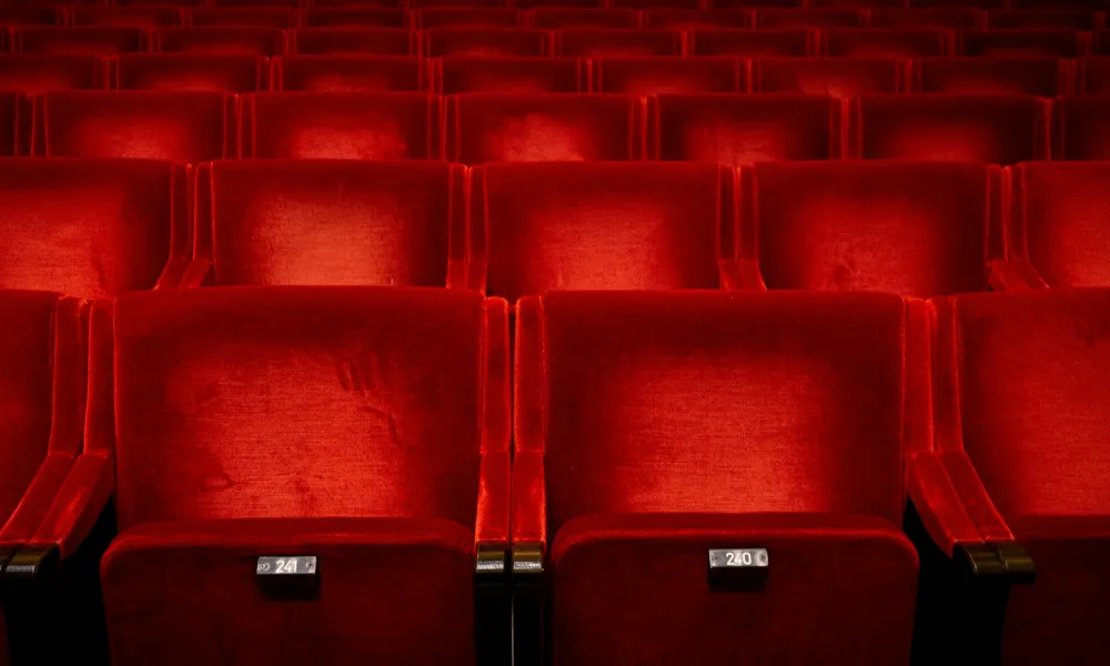 Empty red velvet theater or cinema seats arranged in rows with seat numbers visible on each seat.
