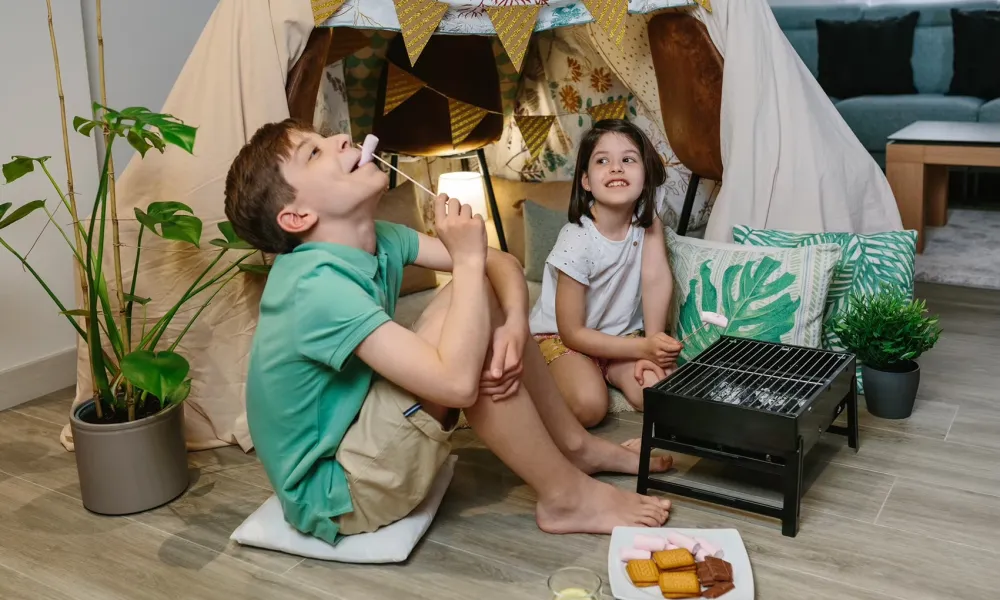 Two children enjoying indoor camping with a blanket tent and roasting marshmallows on a small grill.