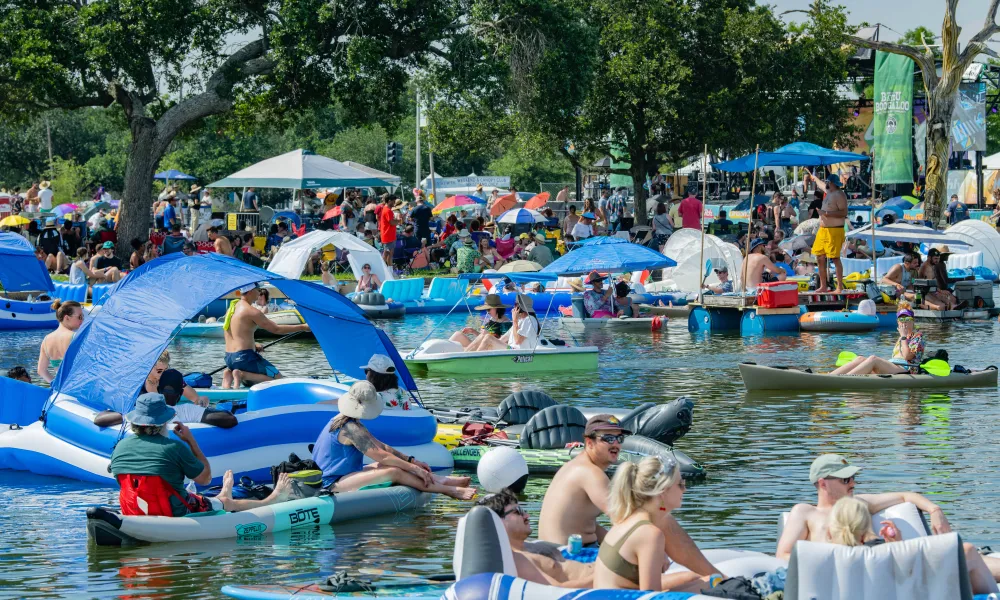 People enjoying a sunny day on inflatable rafts, kayaks, and boats in a crowded lake with trees and tents nearby.