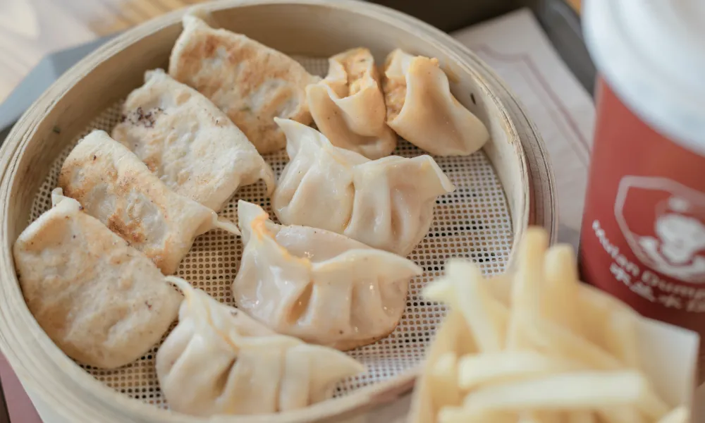Steamed and pan-fried dumplings served in a bamboo basket with French fries and a drink cup nearby
