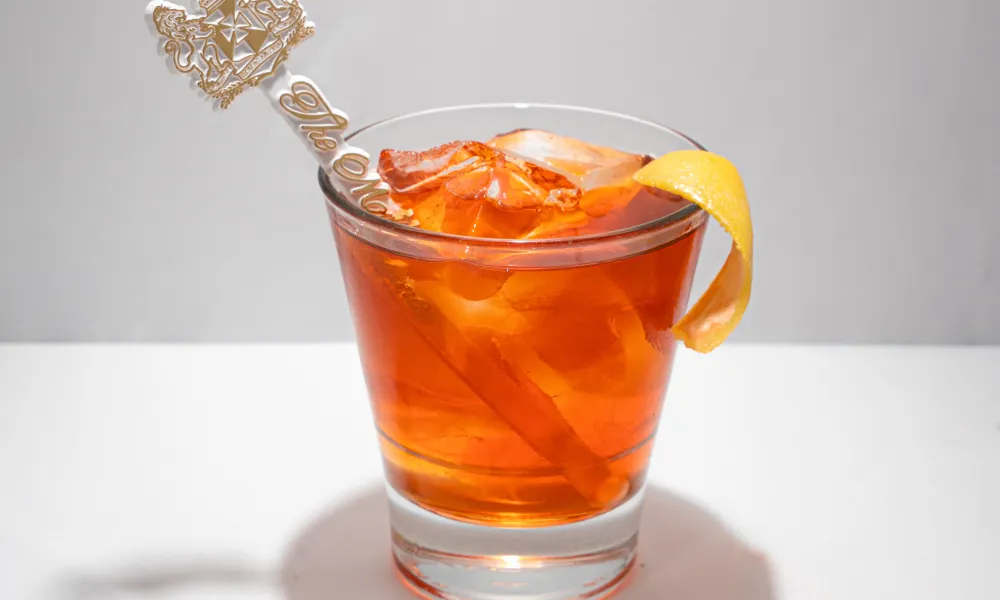 Glass of orange cocktail with ice, lemon twist garnish, and decorative stirrer on a white surface.