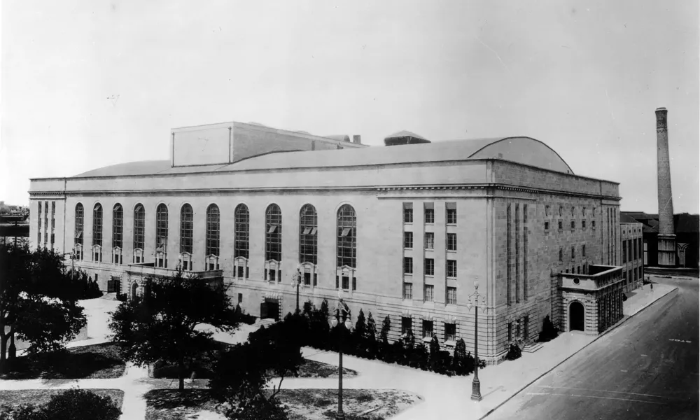 Historic black and white photo of a large classical-style building with tall arched windows and a smokestack in the background.