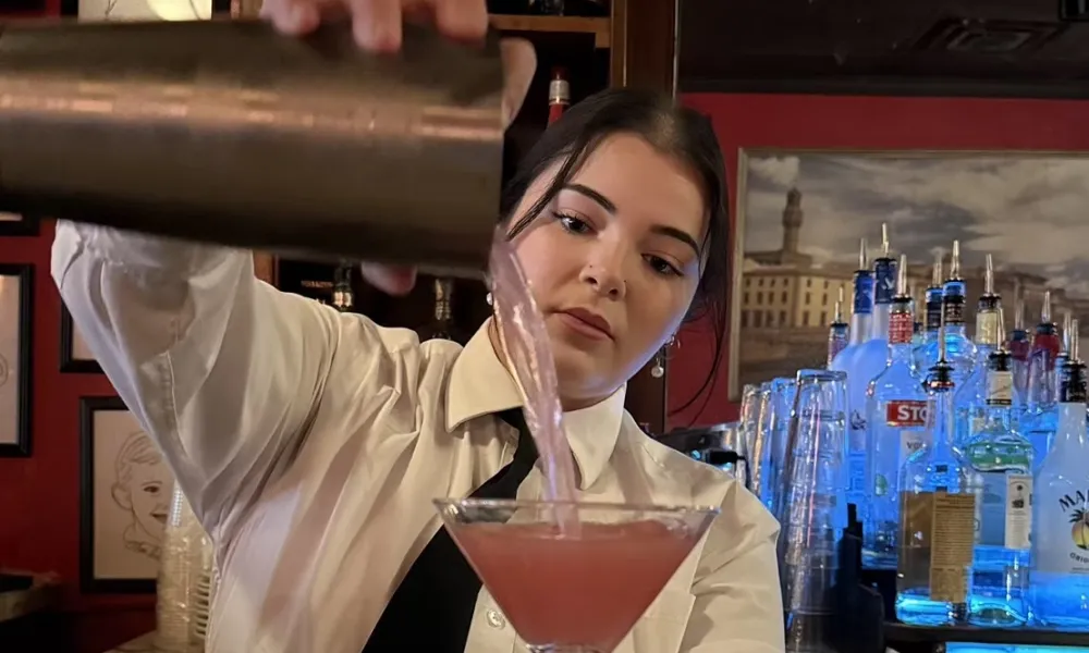Bartender pouring pink cocktail from shaker into martini glass at a bar with bottles and portraits in background