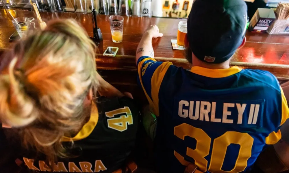 Two sports fans wearing jerseys with Kamara 41 and Gurley II 30 sitting at a bar counter with drinks.