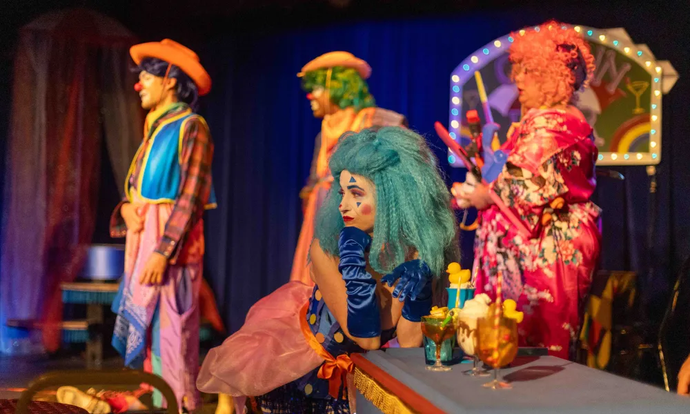 Colorful clowns in vibrant costumes perform on stage with drinks on a table under theatrical lighting.