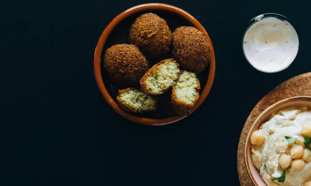 Bowl of golden falafel balls with cut pieces showing inside, served with creamy dip and hummus on black background
