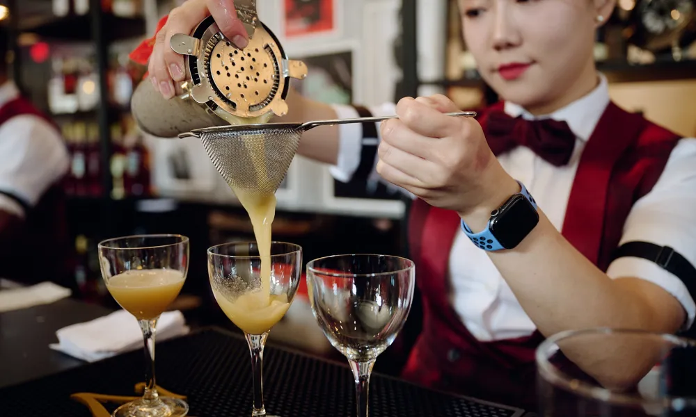 Bartender pouring a cocktail through a strainer into a glass at a bar setting