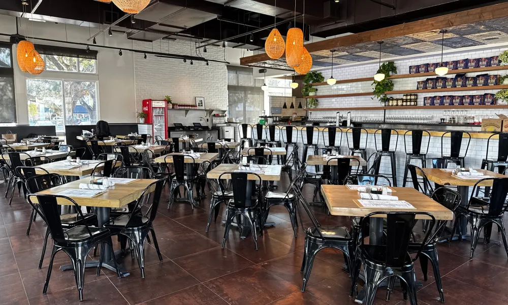 Modern restaurant interior with wooden tables, black metal chairs, pendant lights, and a long bar counter.